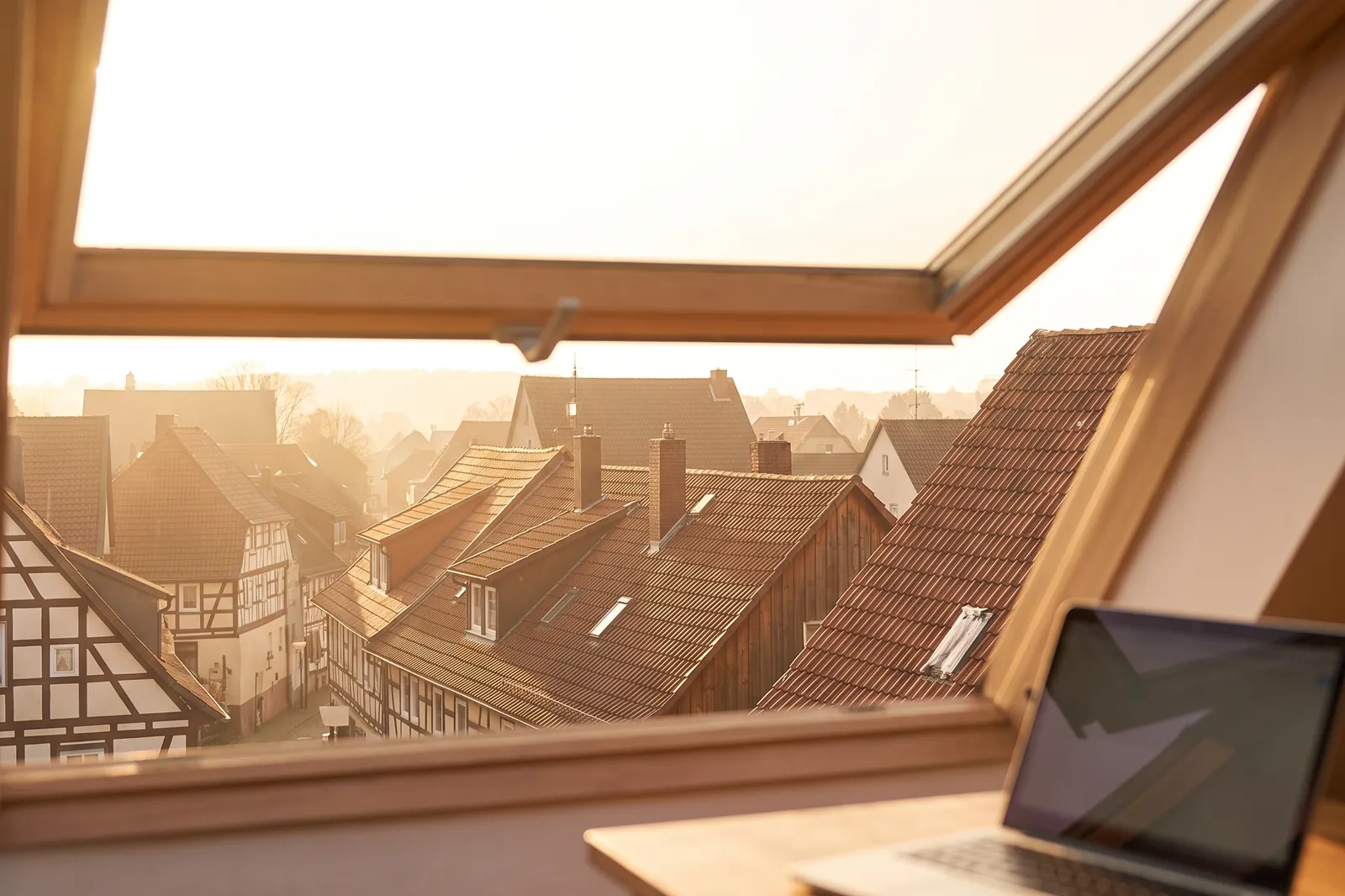 Schreibtisch mit Laptop am geöffneten Dachfenster mit Blick über die Dächer einer Fachwerkstadt