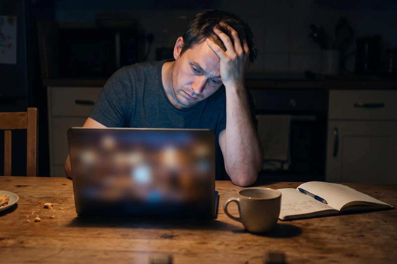 Person sitzt abends allein am Küchentisch vor dem Laptop und stützt den Kopf in die Hand